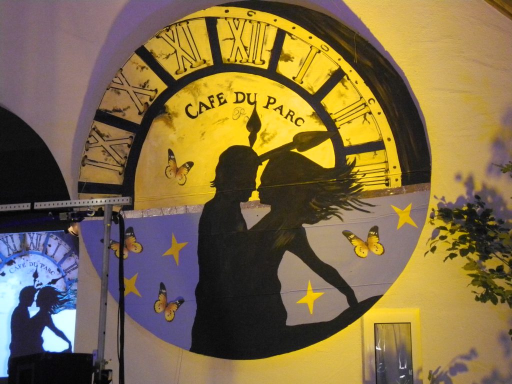 riesiges Zifferblatt mit der Aufschrift "Café du Parc". Im Vordergrund die Silhouette eines tanzenden Paares